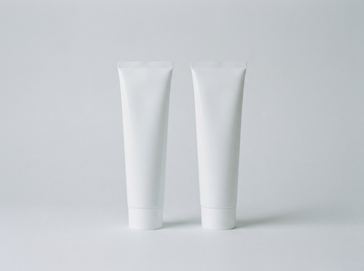 Two plain unmarked white toothpaste tubes standing side by side on a pale grey background