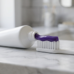 Purple toothpaste squeezed from a plain white tube onto a white toothbrush on a marble bathroom counter